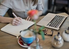 Person Writing On A Notebook Beside Macbook
